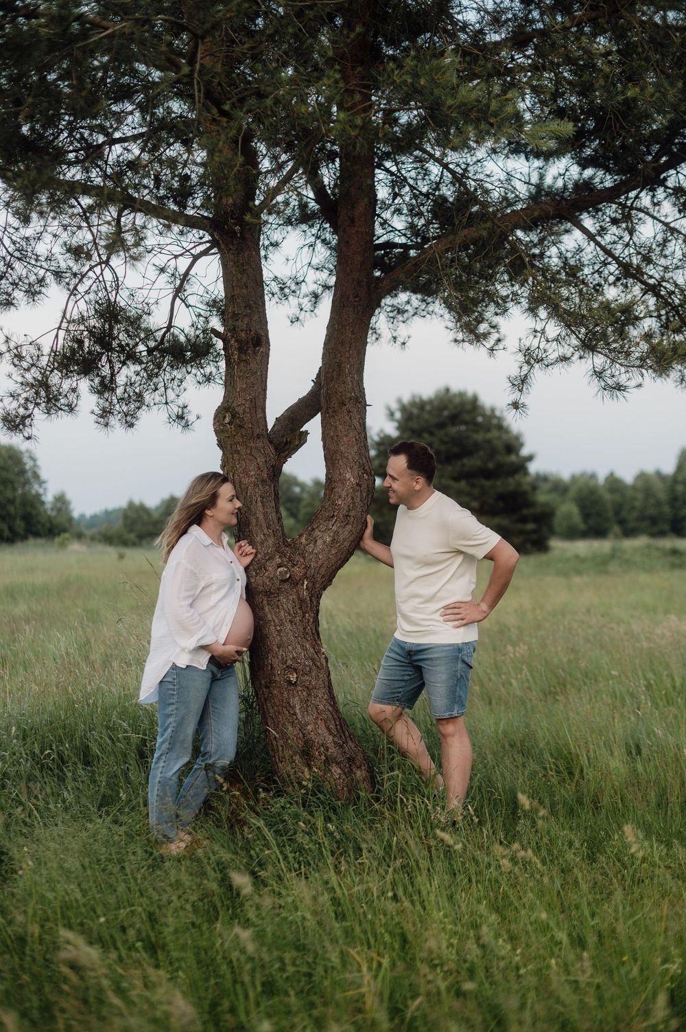 A couple posing playfully by a large tree in a grassy field on a cloudy day.