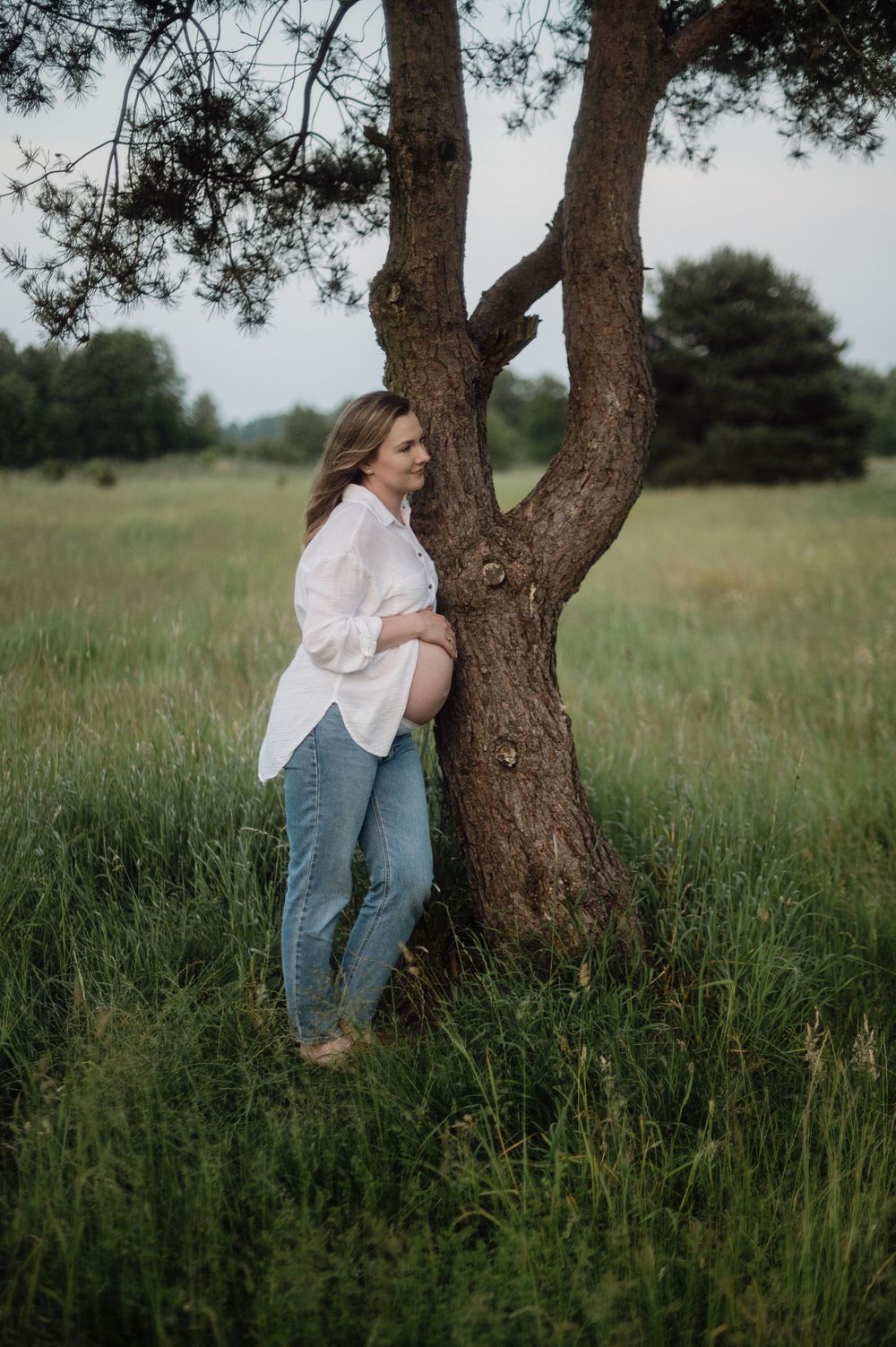 Maternity portrait of someone leaning against a forked tree trunk in a grassy field.