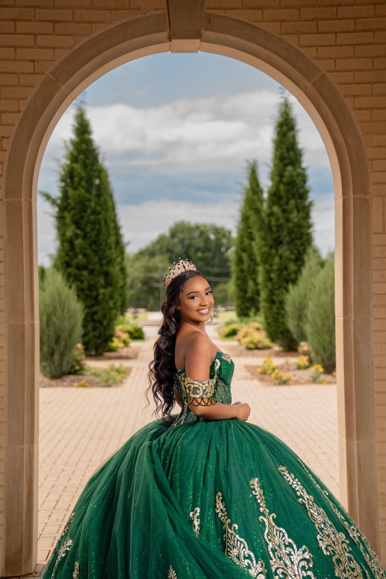 A person wearing a champagne-colored ballgown with floral appliques poses in a sunlit garden setting.