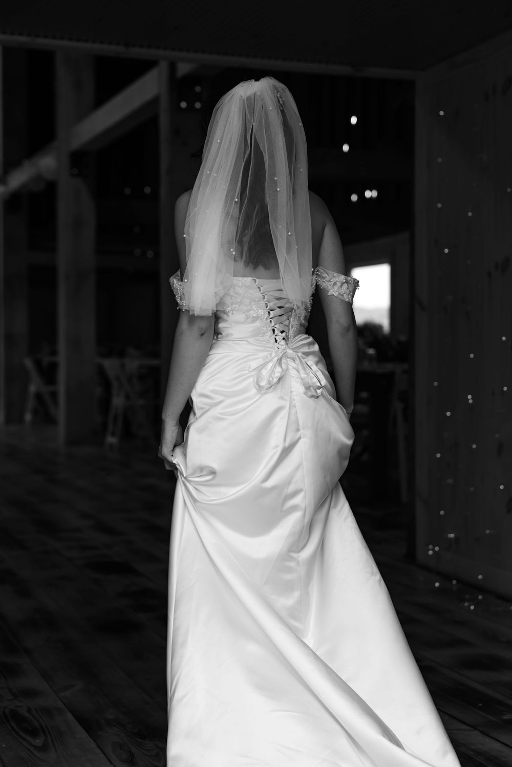 Black and white rear view of bride in elegant lace corset gown and veil standing in barn doorway.