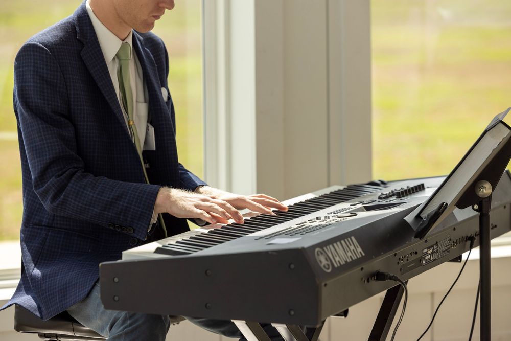 Man in blue suit playing a Yamaha keyboard at a wedding reception near large windows.