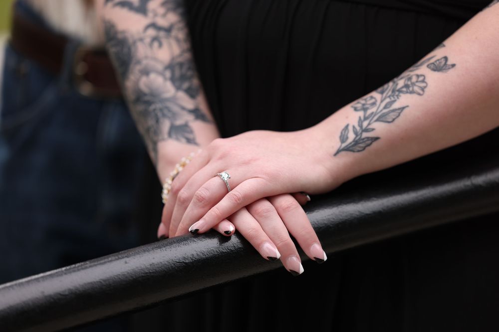Close-up of two tattooed arms resting on a black railing, with an engagement ring visible on one hand.