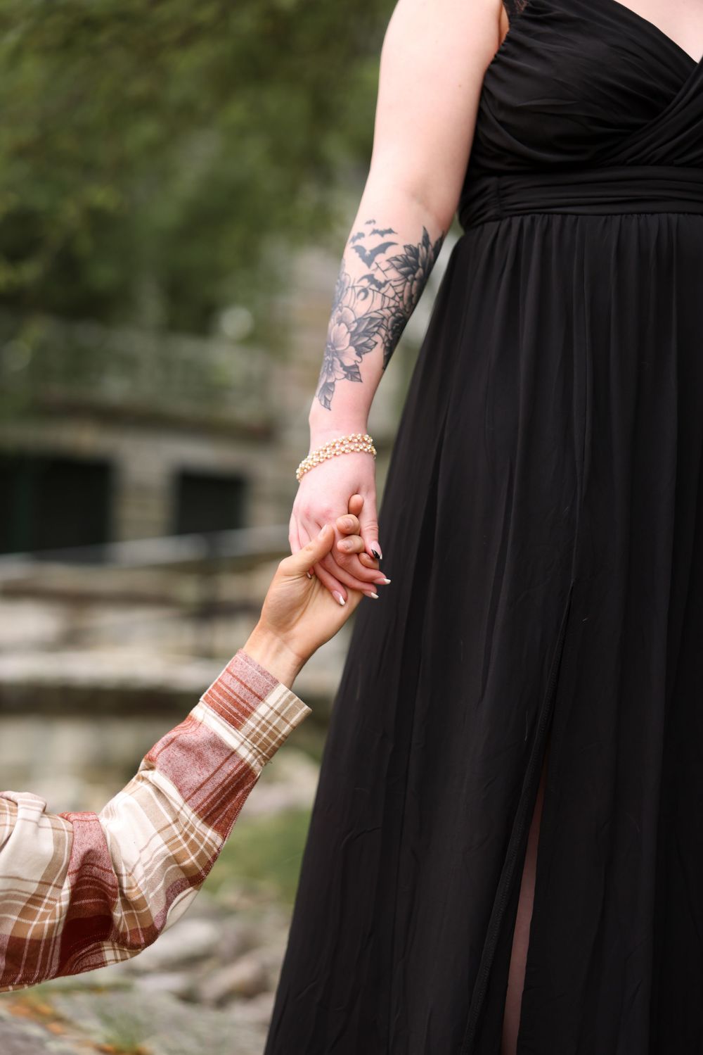 A woman in a black dress with floral arm tattoos holds hands with a man in a plaid shirt outdoors.