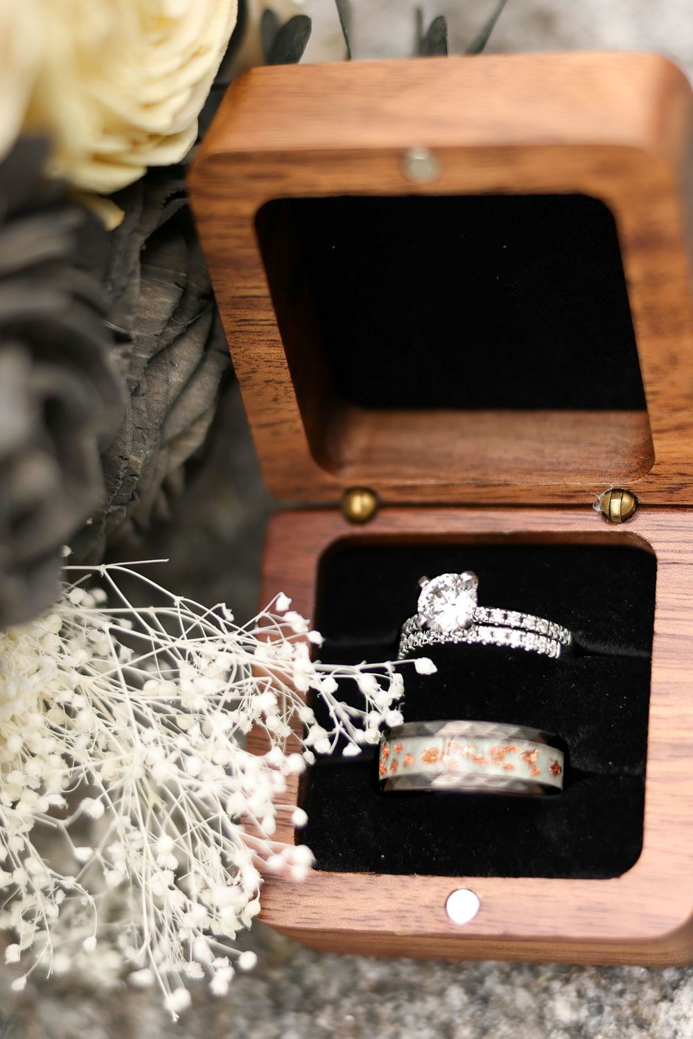 Wedding rings displayed in an open wooden ring box with black velvet lining, surrounded by flowers.
