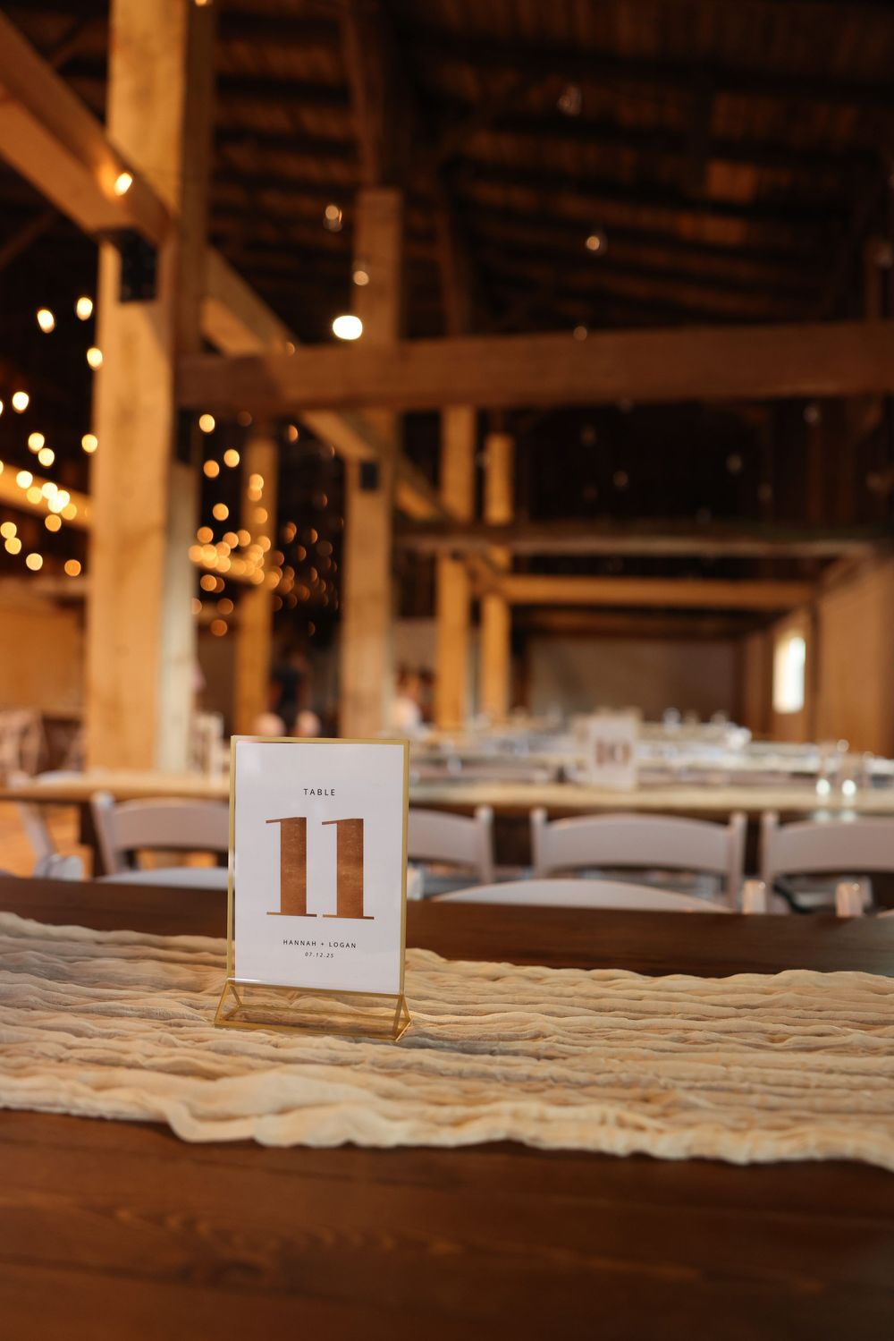Wedding reception table number 11 card in gold holder on linen runner inside a warmly lit barn venue.