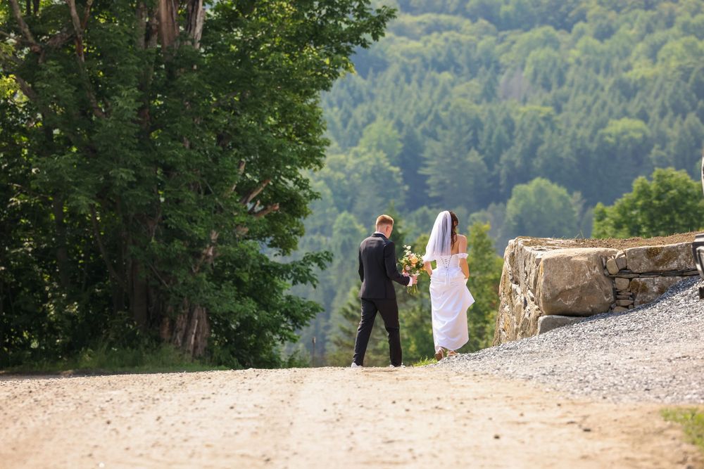Bride and groom walking hand in hand along a gravel path surrounded by lush Vermont forest scenery.