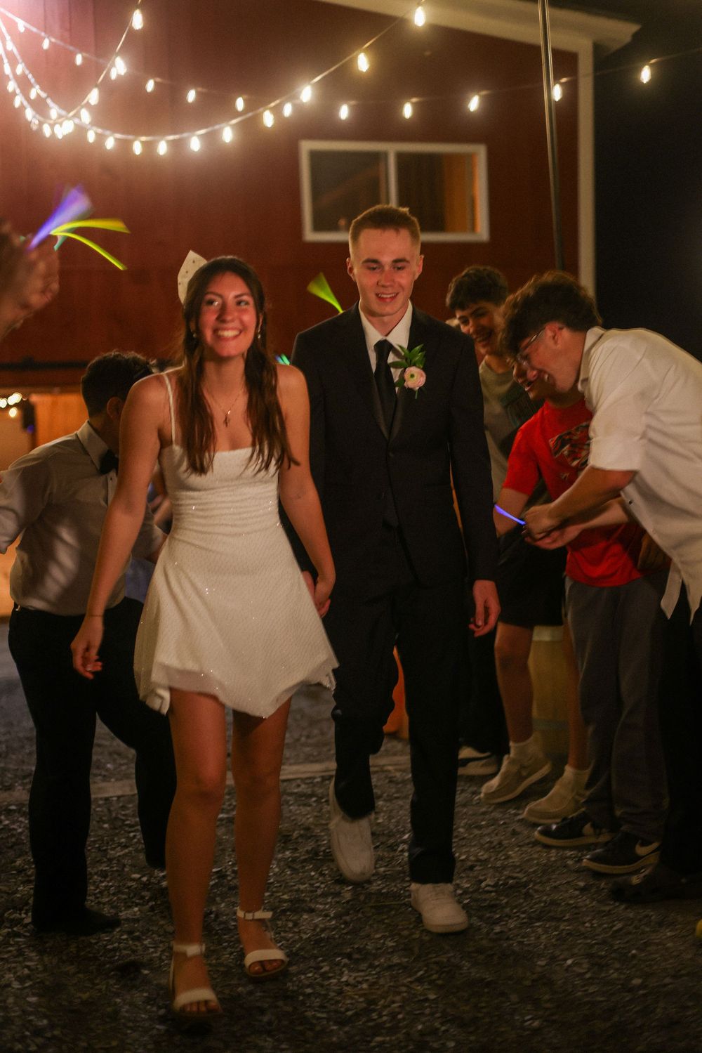 Smiling bride and groom walking through a celebratory send-off line with glow sticks at night.