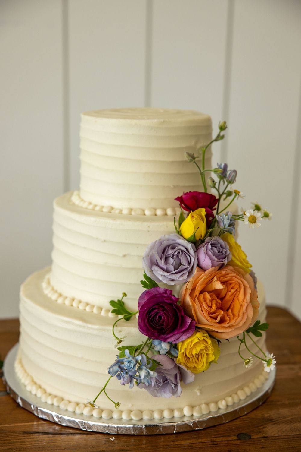 Three-tier white wedding cake with colorful fresh flower arrangement on wooden table.