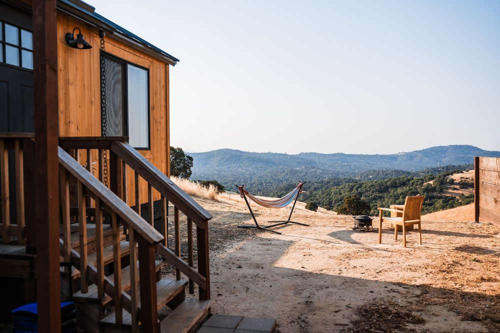 A wooden cabin with a hammock and chairs overlooks mountains on a rustic hilltop property.