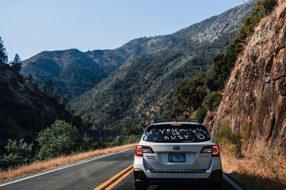 A silver Subaru Outback drives along a scenic mountain road with rugged cliffs and forest views.