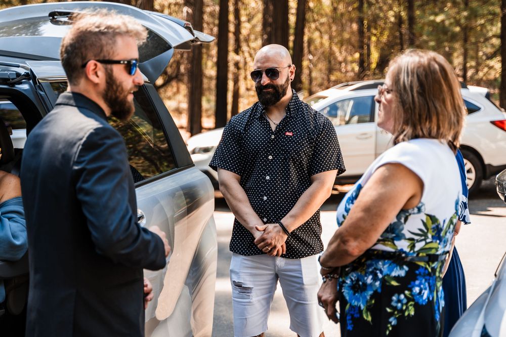 Three people having a conversation in a parking area surrounded by trees and parked vehicles.