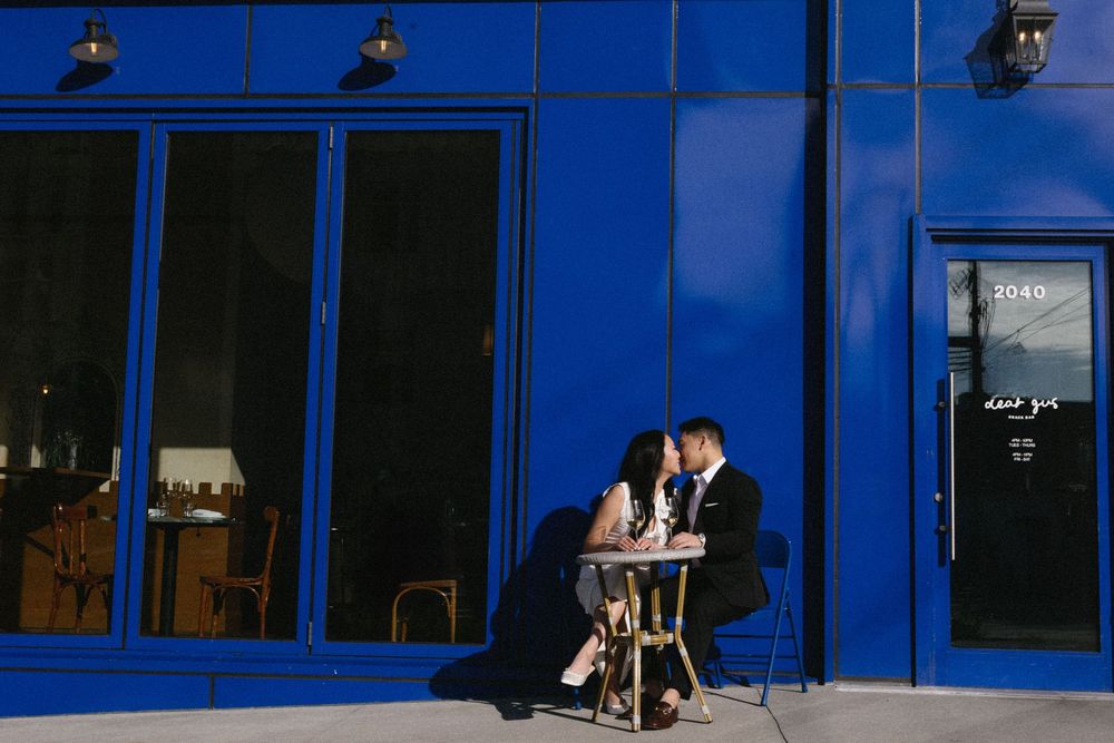 A couple shares a romantic moment at a table outside a blue-painted storefront.