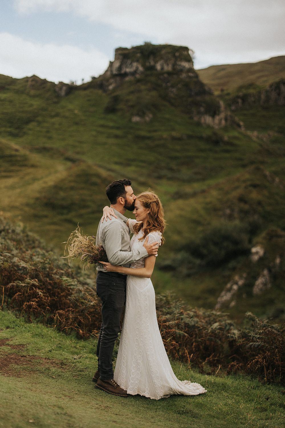 Couple shares romantic embrace on green hillside with dramatic cliffs and mountains in Scottish highlands landscape.