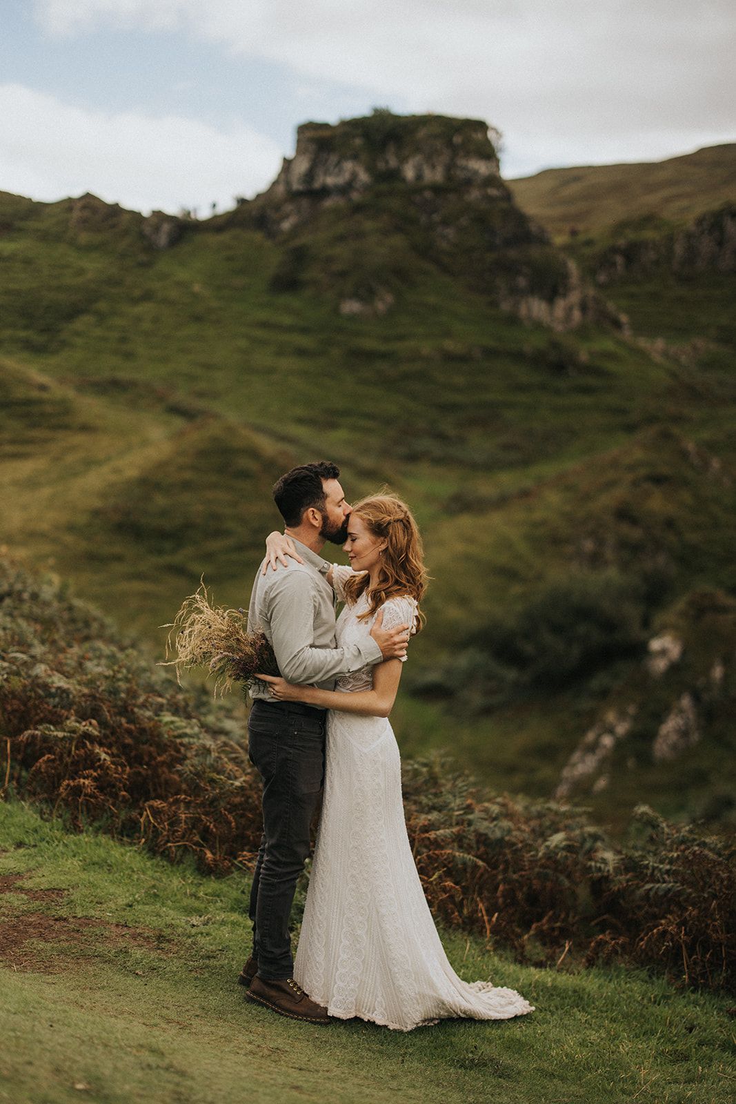 Couple shares romantic embrace on green hillside with dramatic cliffs and mountains in Scottish highlands landscape.