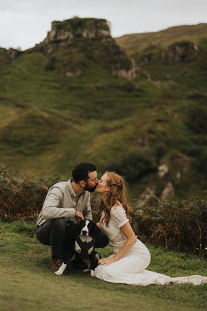 A romantic couple sits with their border collie dog on a grassy hillside with dramatic rocky cliffs in the background.