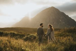 Two figures hold hands while standing in a golden meadow at sunset with a majestic mountain silhouetted in the background.