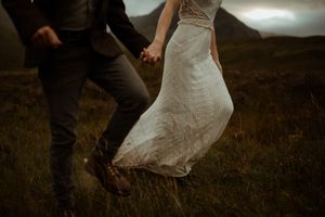 A couple walks hand in hand through a grassy meadow at sunset wearing wedding attire.