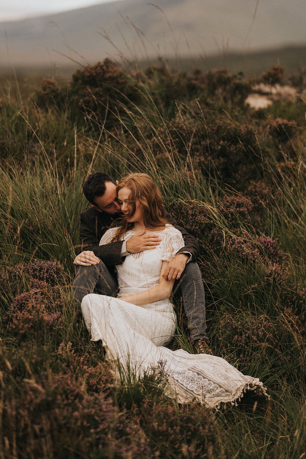 Romantic couple sit nestled together on a hillside of purple heather during sunset in an intimate outdoor moment.
