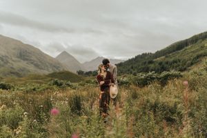 A couple shares an intimate moment on a hiking trail surrounded by misty Scottish mountains and wildflowers.