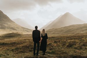 Two figures stand together overlooking a misty mountain landscape in the Scottish Highlands on an overcast day.