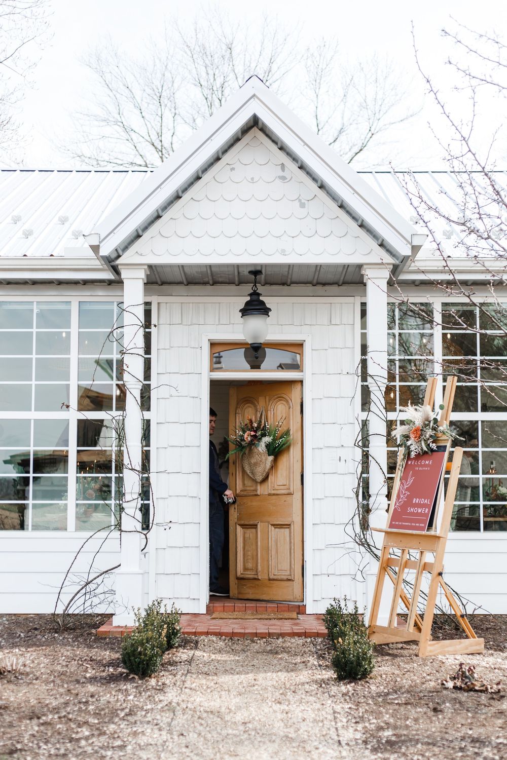 White brick farmhouse entryway with wooden door, holiday wreath, flanking windows, and rustic wood easel on the porch.