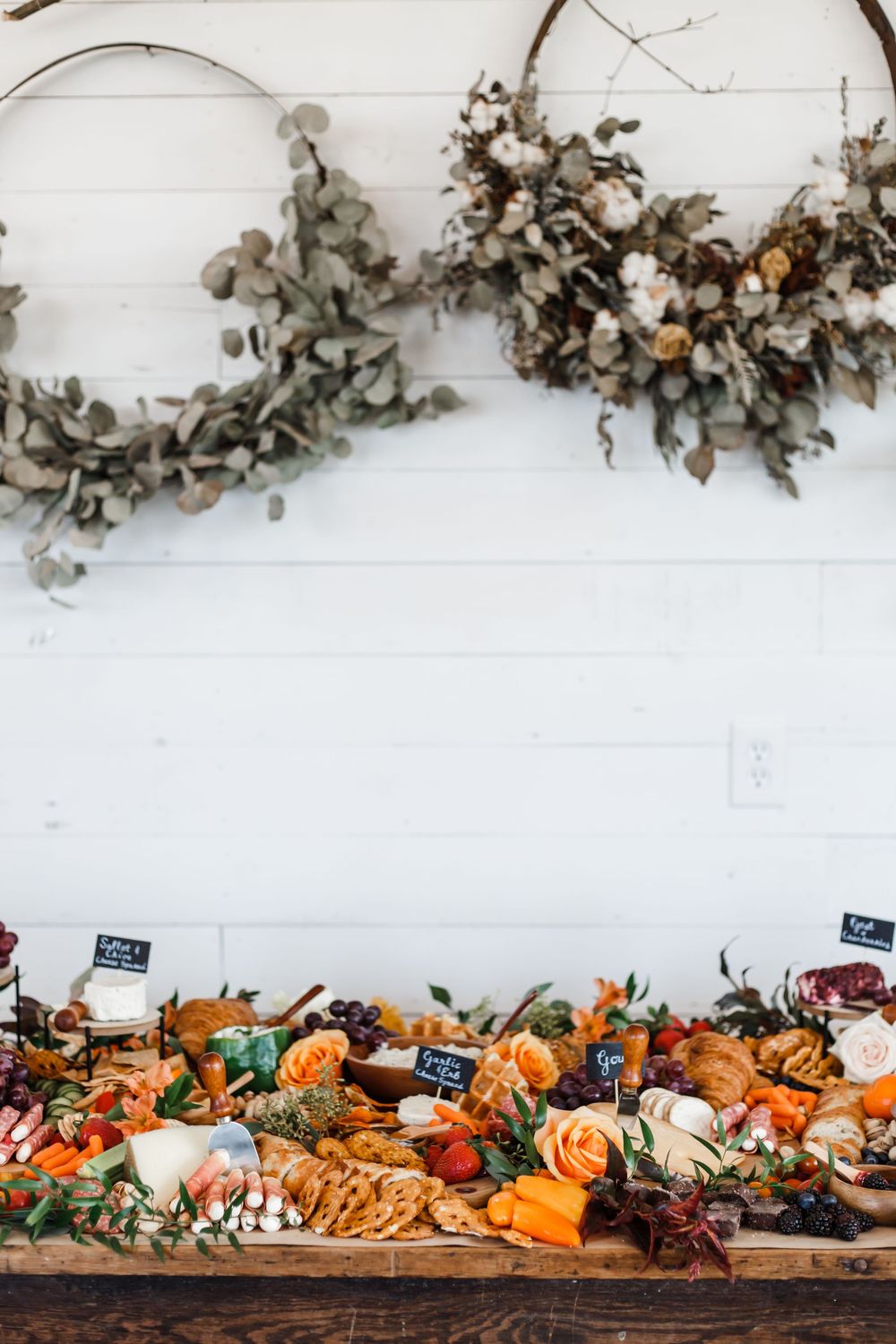 Rustic grazing table with dried florals hanging above featuring assorted meats, cheeses and seasonal fruits.