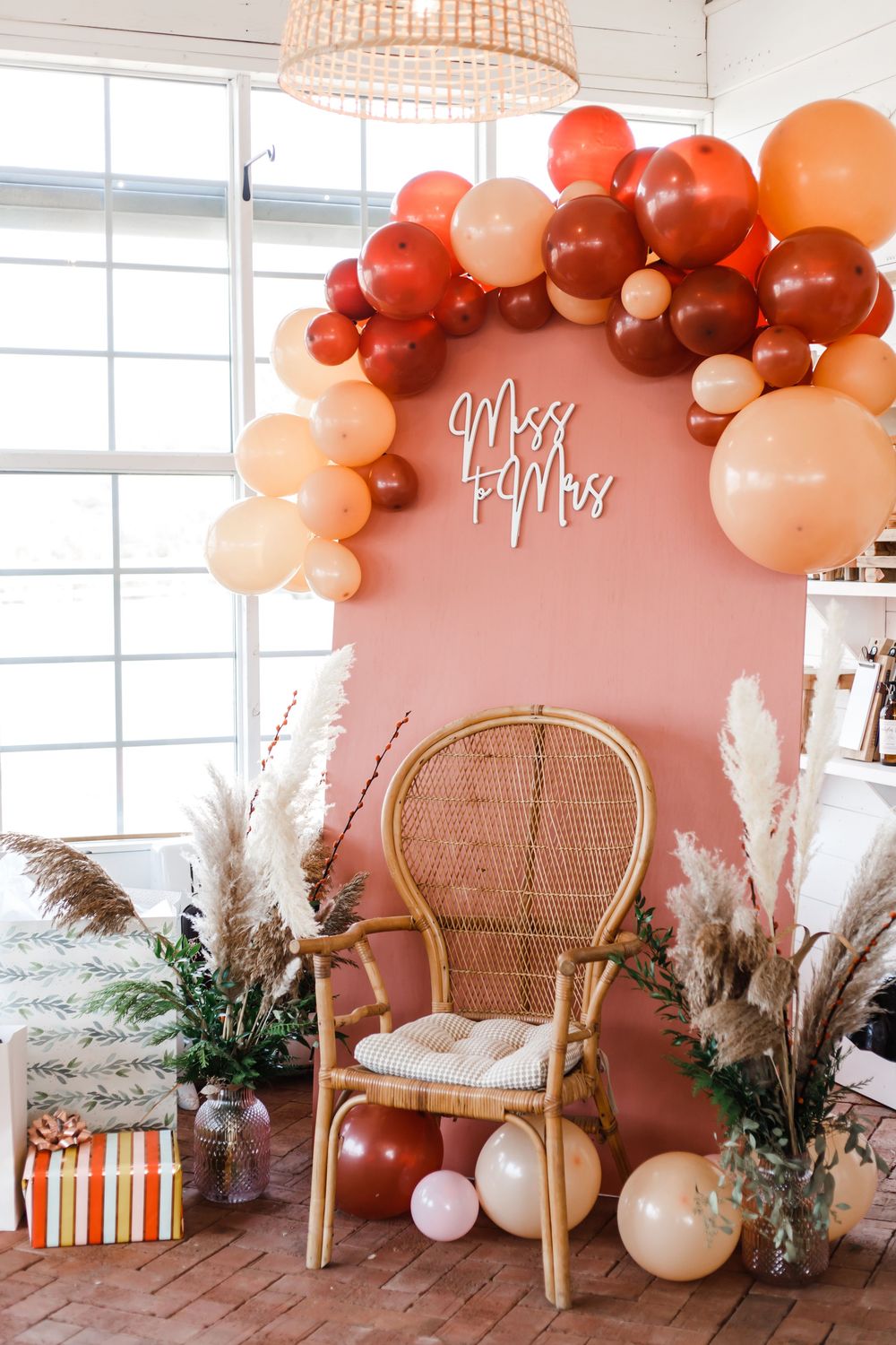 Boho-inspired event setup with peachy balloon arch, rattan chairs, and pampas grass against white windows.