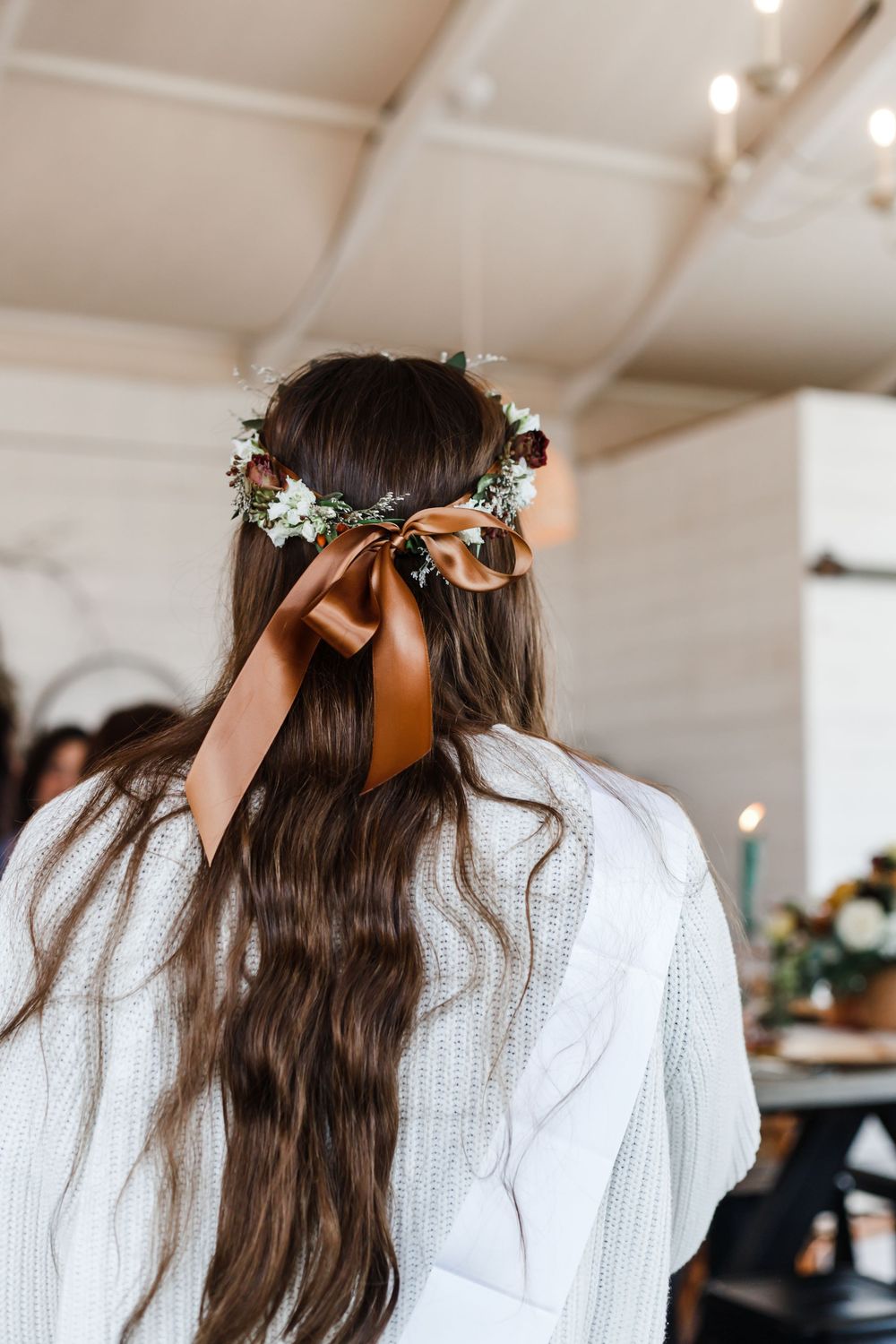 Long brown hair adorned with floral crown and copper ribbon against white interior setting.