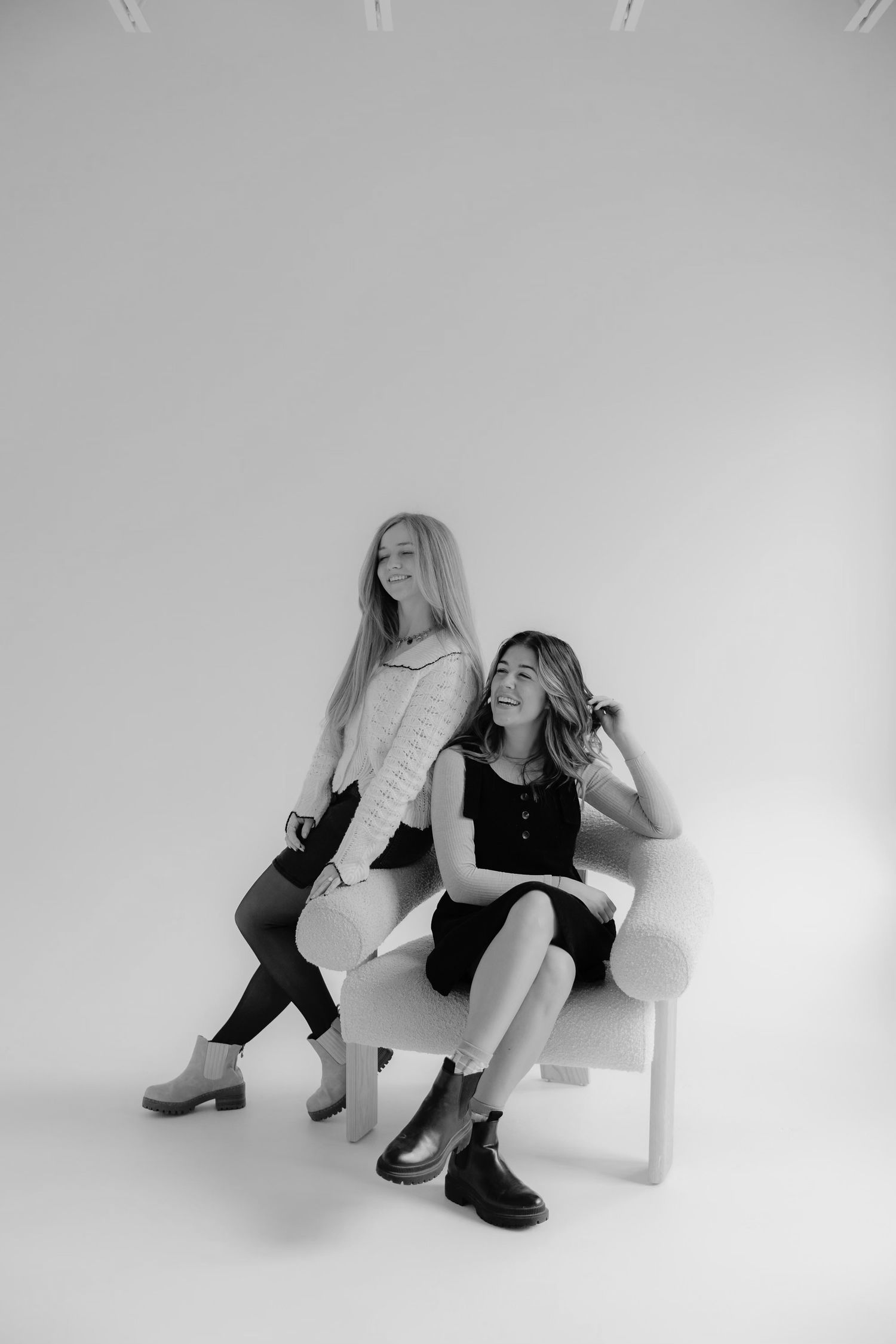 Black and white fashion portrait of two models sitting casually on white bench in studio setting.