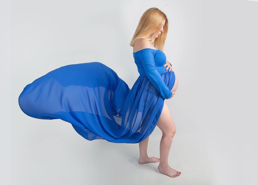 Artistic maternity session with flowing royal blue dress against white background.