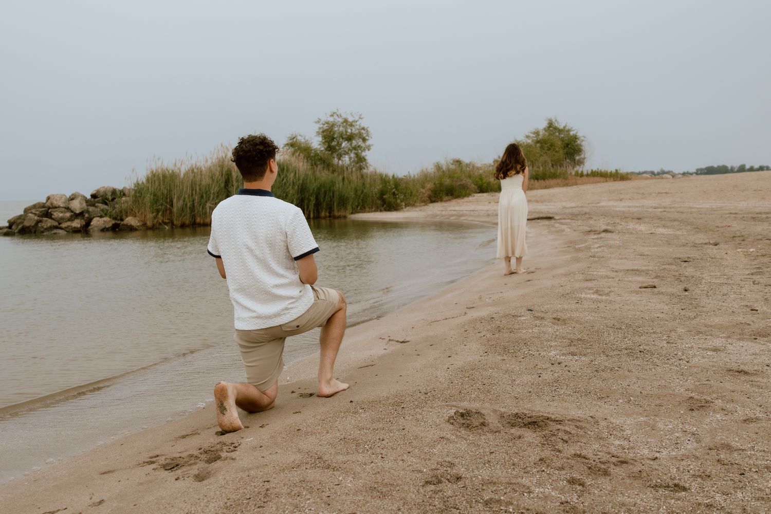Couple on a sandy beach shore on an overcast day, woman standing near the water.