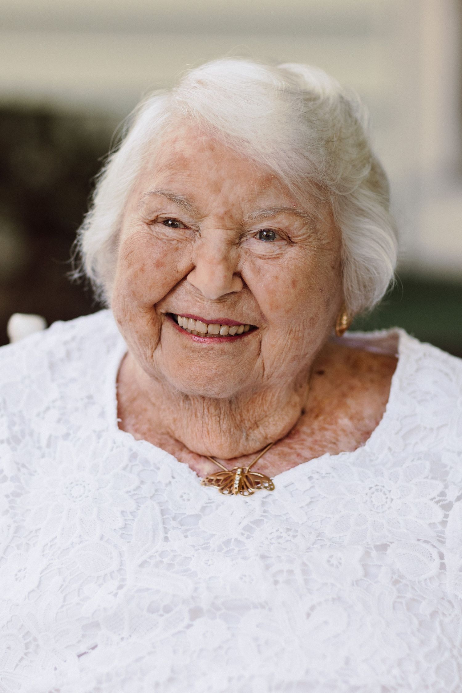 Elderly person wearing white lace dress and necklace smiling warmly at camera.