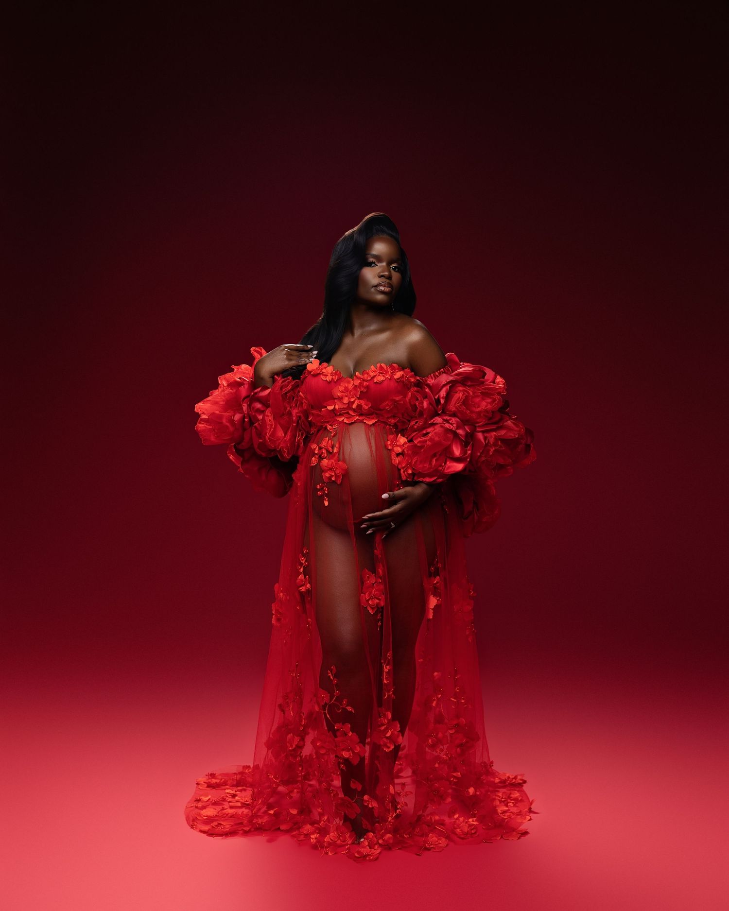 Pregnant woman in elegant red floral sheer robe posing gracefully against a red studio background.