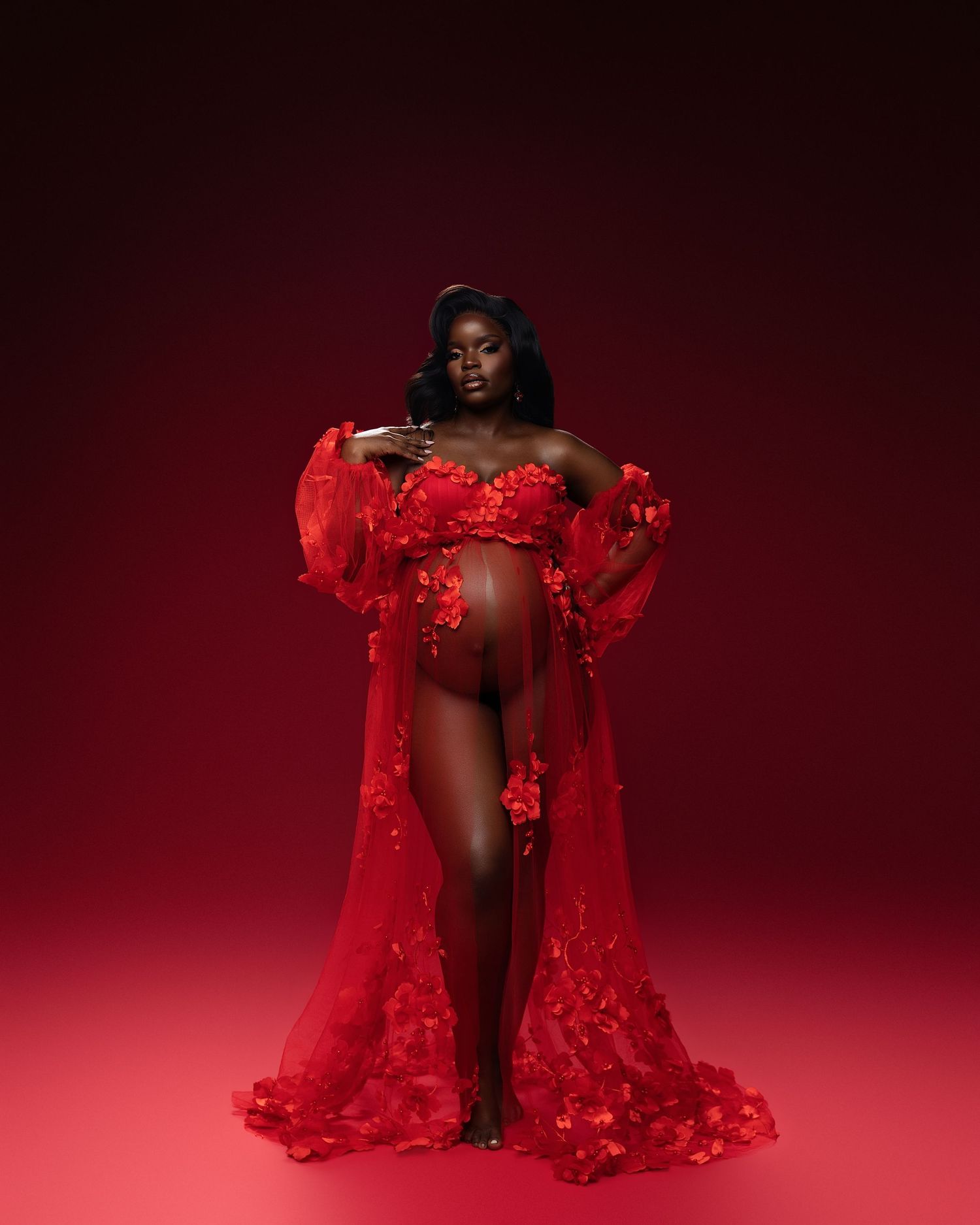 Pregnant woman in red floral sheer maternity gown posing against a red studio background.