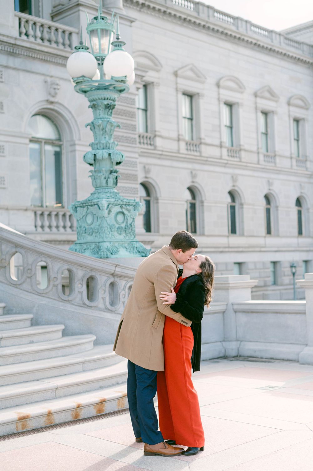 Couple embracing in front of classical white building with ornate turquoise lampposts.