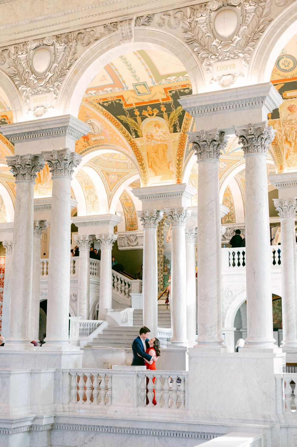 Couple embraces in grand hall with white columns and gilded ceiling details.