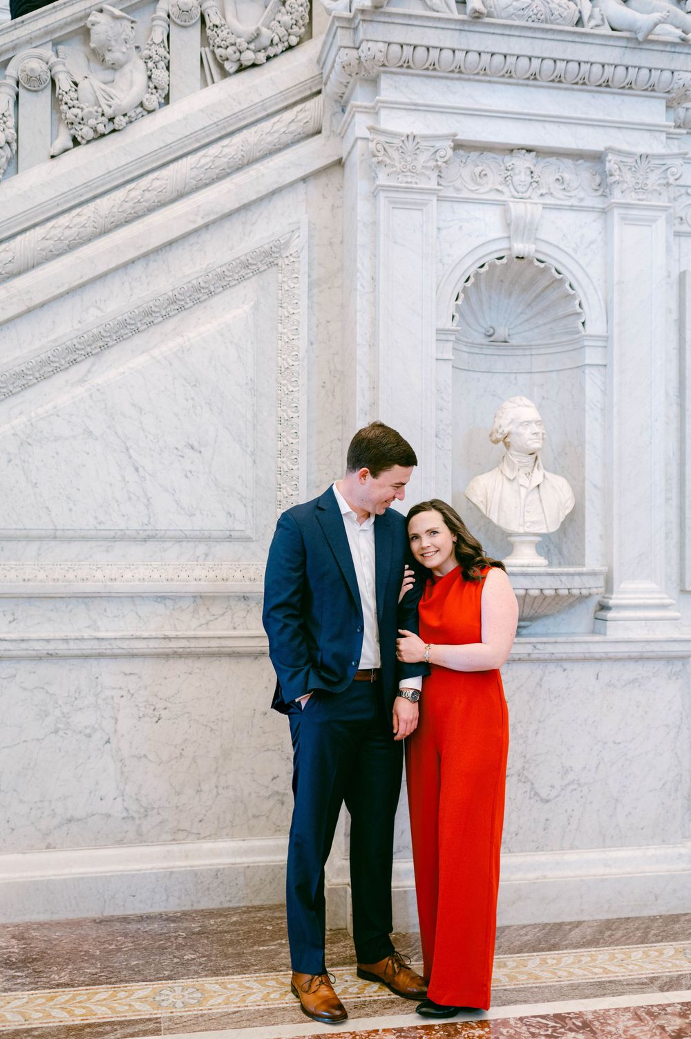 Romantic couple poses together against ornate white marble walls in elegant formal attire.