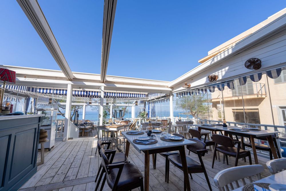 A modern outdoor rooftop dining area with wooden tables and chairs overlooking a cityscape under white awnings.