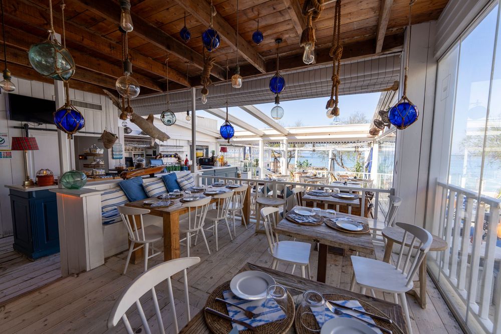Bright waterfront dining room with wooden beams, blue pendant lights, and white furniture overlooking the ocean.
