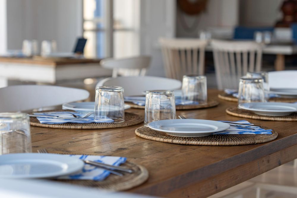 Place settings with blue napkins and glass tumblers arranged on a rustic wooden dining table in natural light.