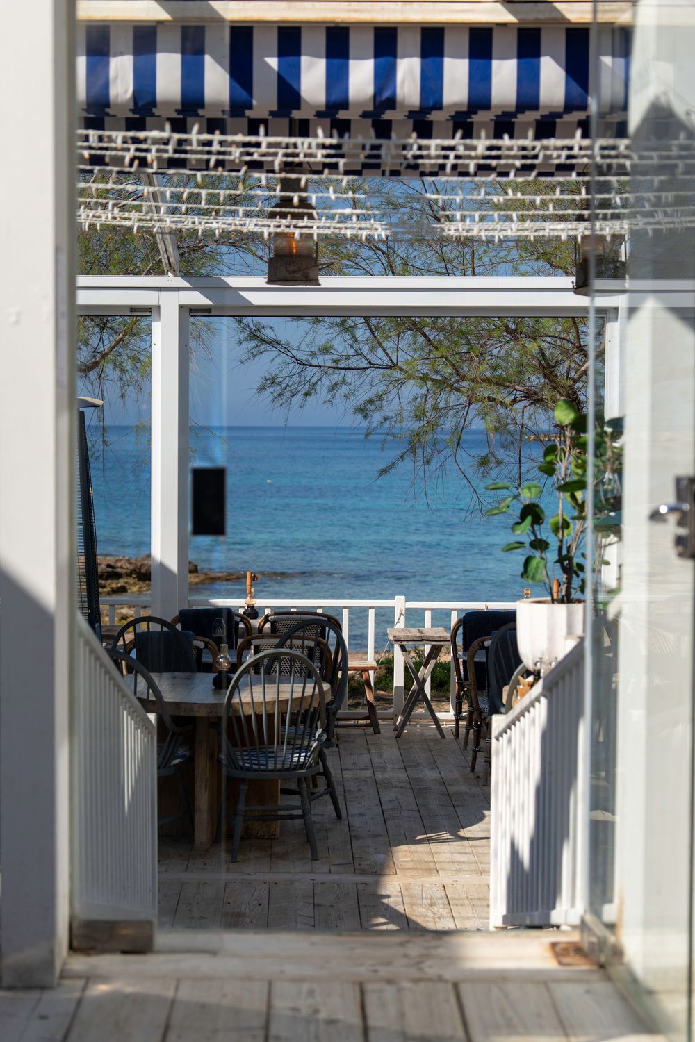 View through doorway of seaside restaurant patio with wooden chairs and striped awning overlooking turquoise ocean waters.