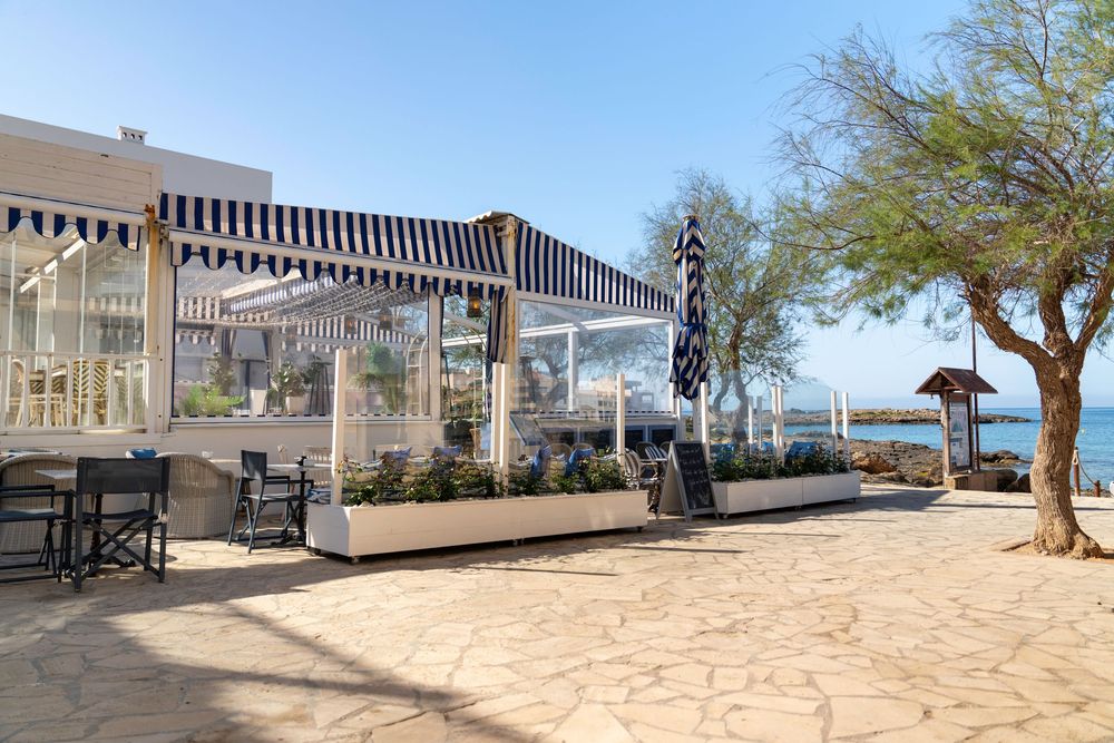 Outdoor beachfront restaurant terrace with modern pergola, wooden furniture and desert landscaping under sunny skies.