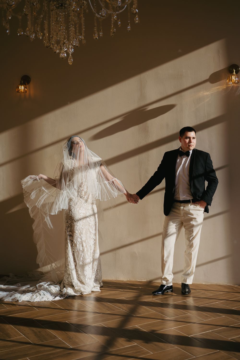 Bride in lace gown and veil holding hands with groom under a chandelier at Hotel Nangi, Valle de Guadalupe.
