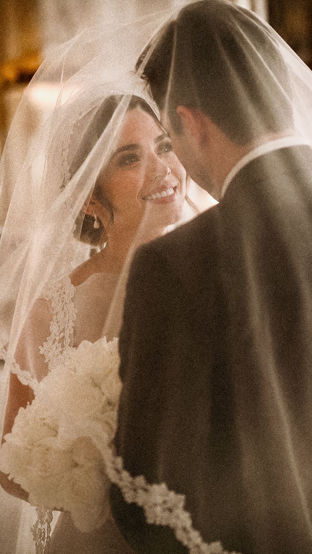 Smiling bride under a lace veil gazing at her groom during a romantic wedding ceremony in Valle de Guadalupe, Baja California.