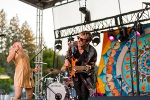 Musicians perform on stage at outdoor festival with colorful mandala backdrop and stage lighting equipment.