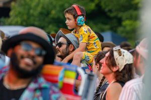Festival attendees watch a performance while a small child wearing headphones sits on someone's shoulders.