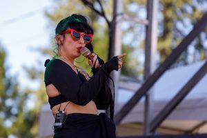 A performer in dark attire and sunglasses sings into a microphone at an outdoor event.