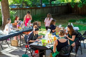 People gather at picnic tables in a shady outdoor area for a summer community meal together.