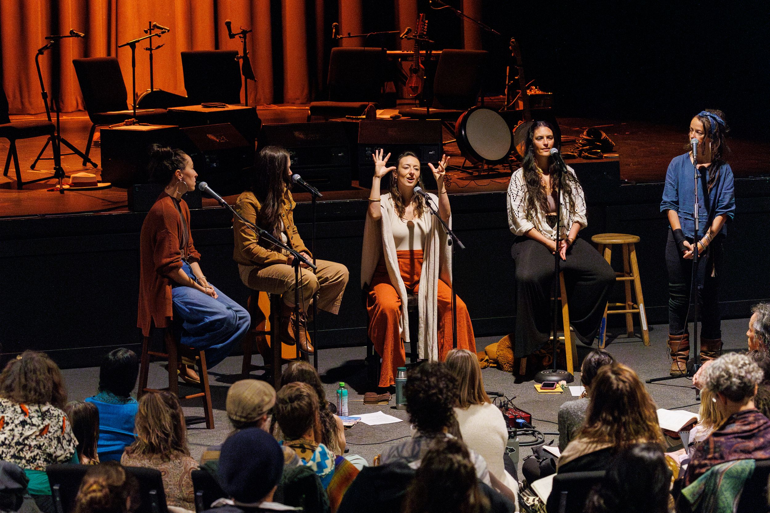 Musicians perform on a wooden stage with stools at a folk concert while audience members watch from below.