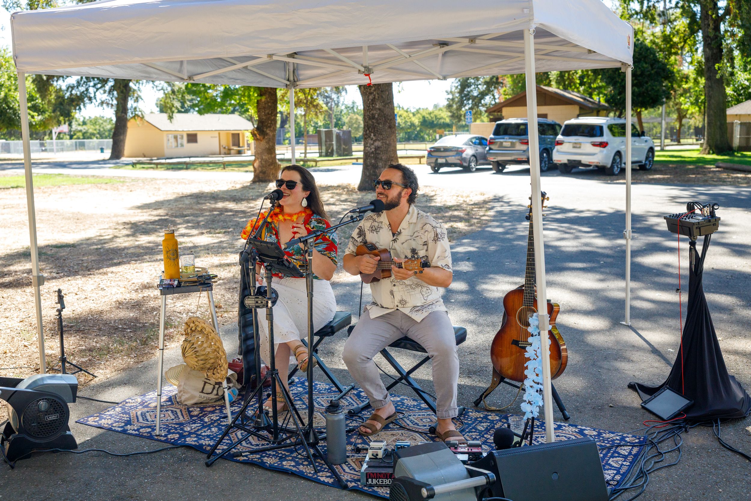 Musicians perform outdoors under a white canopy tent with various instruments and equipment set up on a sunny day.